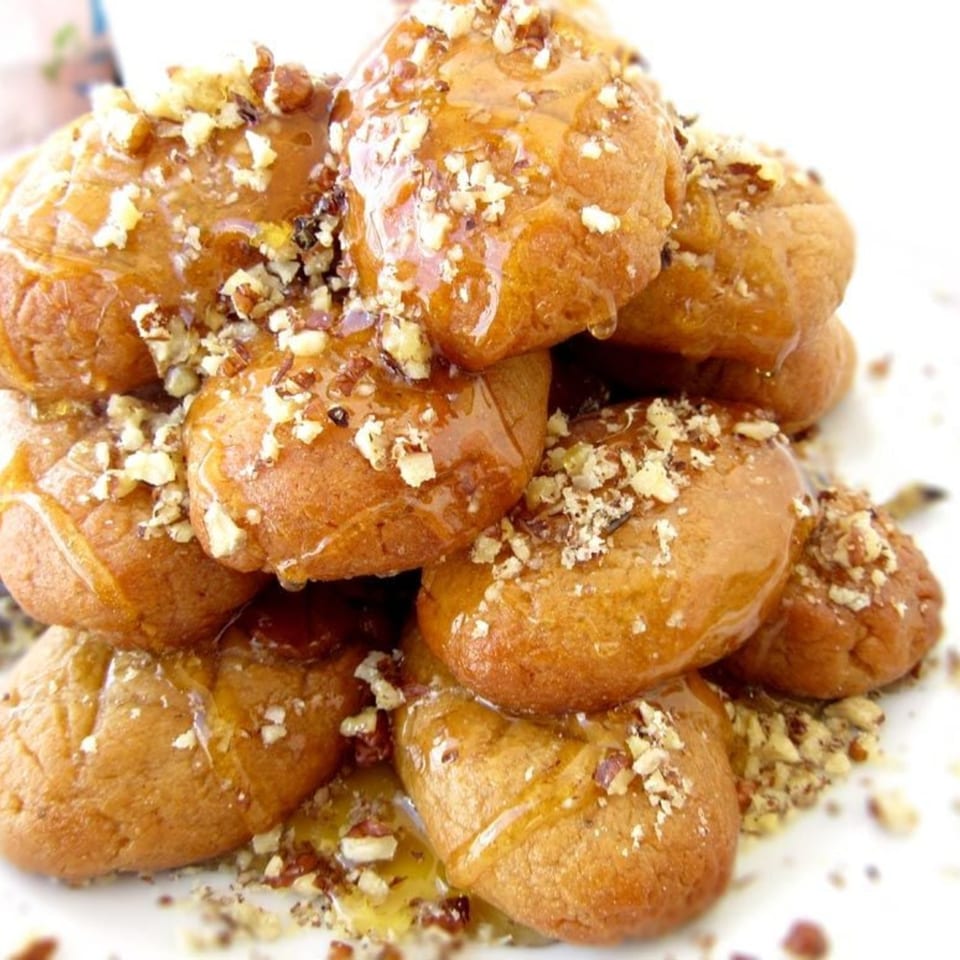A close-up of golden-brown Greek honey cookies (melomakarona) drizzled with honey and topped with chopped walnuts.