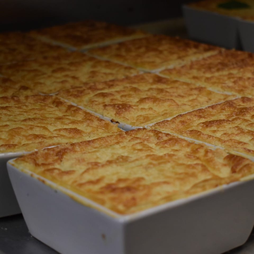 Multiple rectangular dishes of golden-brown, baked Greek pastitsio or moussaka with a creamy topping.