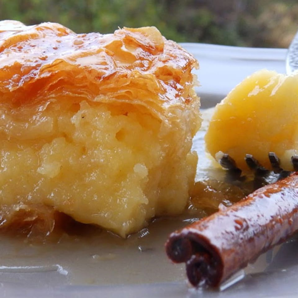 Close-up of a golden Greek dessert, possibly baklava, with a cinnamon stick and fruit.