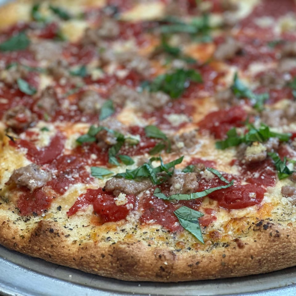 Close-up of a Greek pizza with sausage, tomato sauce, cheese, and fresh basil on a metal tray.