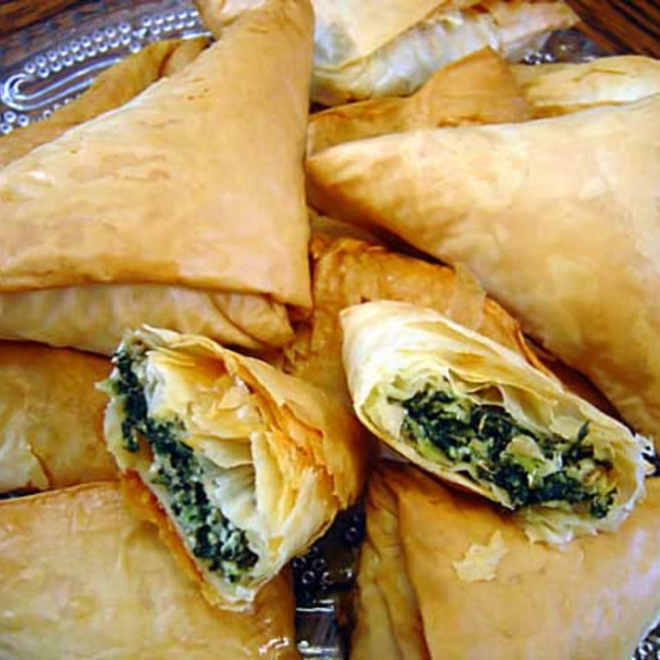 A close-up of golden-brown Greek spanakopita pastries, some cut open revealing spinach and feta filling.