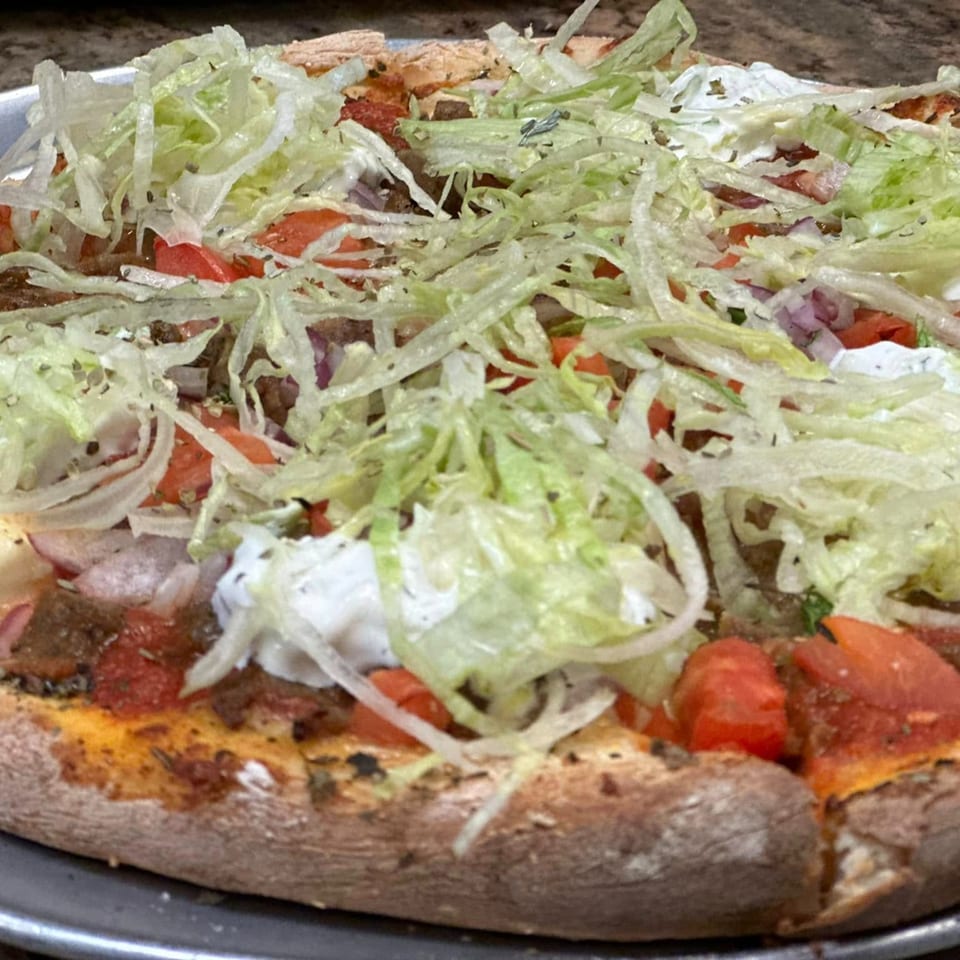 Close-up of a Greek pizza topped with shredded lettuce, diced tomatoes, onions, and white sauce.