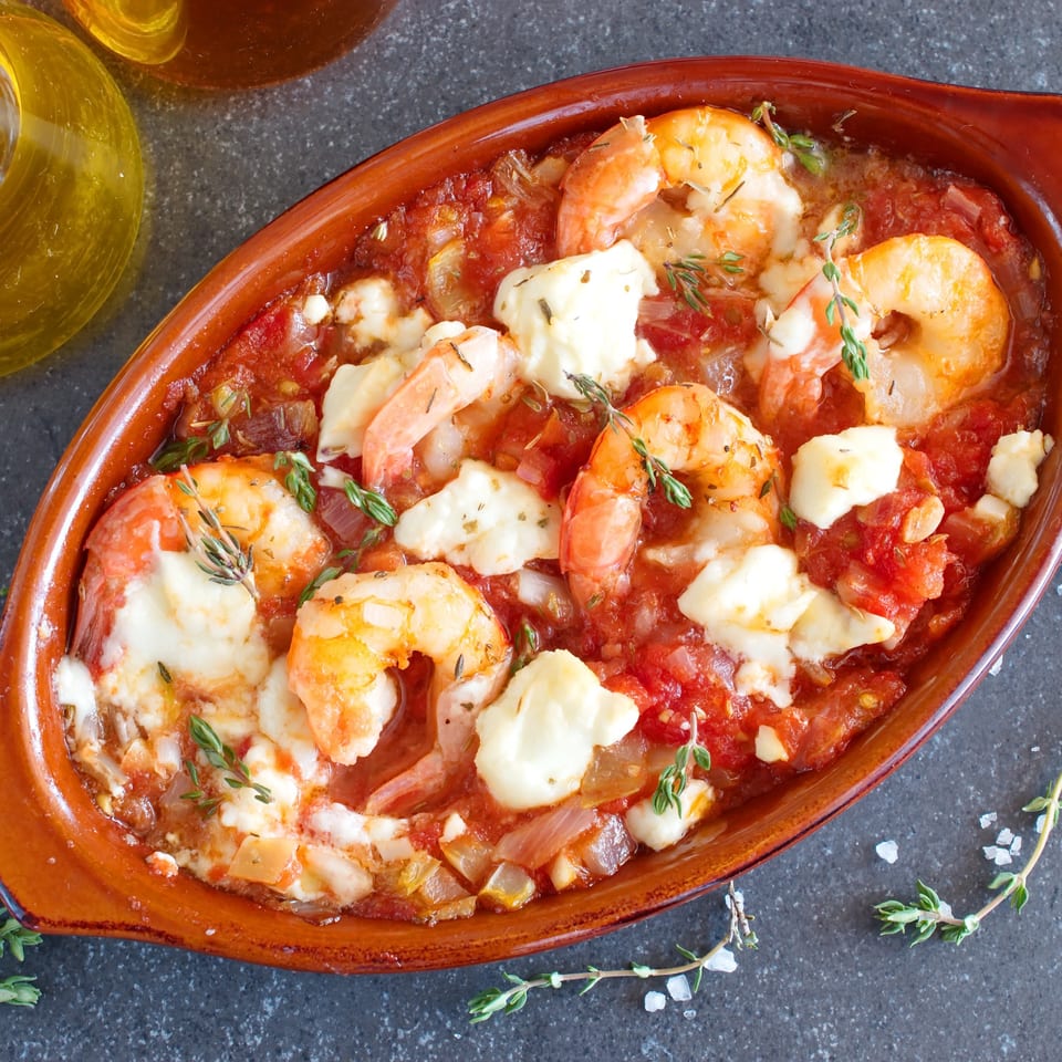 Saganaki shrimp with feta cheese, tomato sauce, and fresh herbs in a brown ceramic dish.