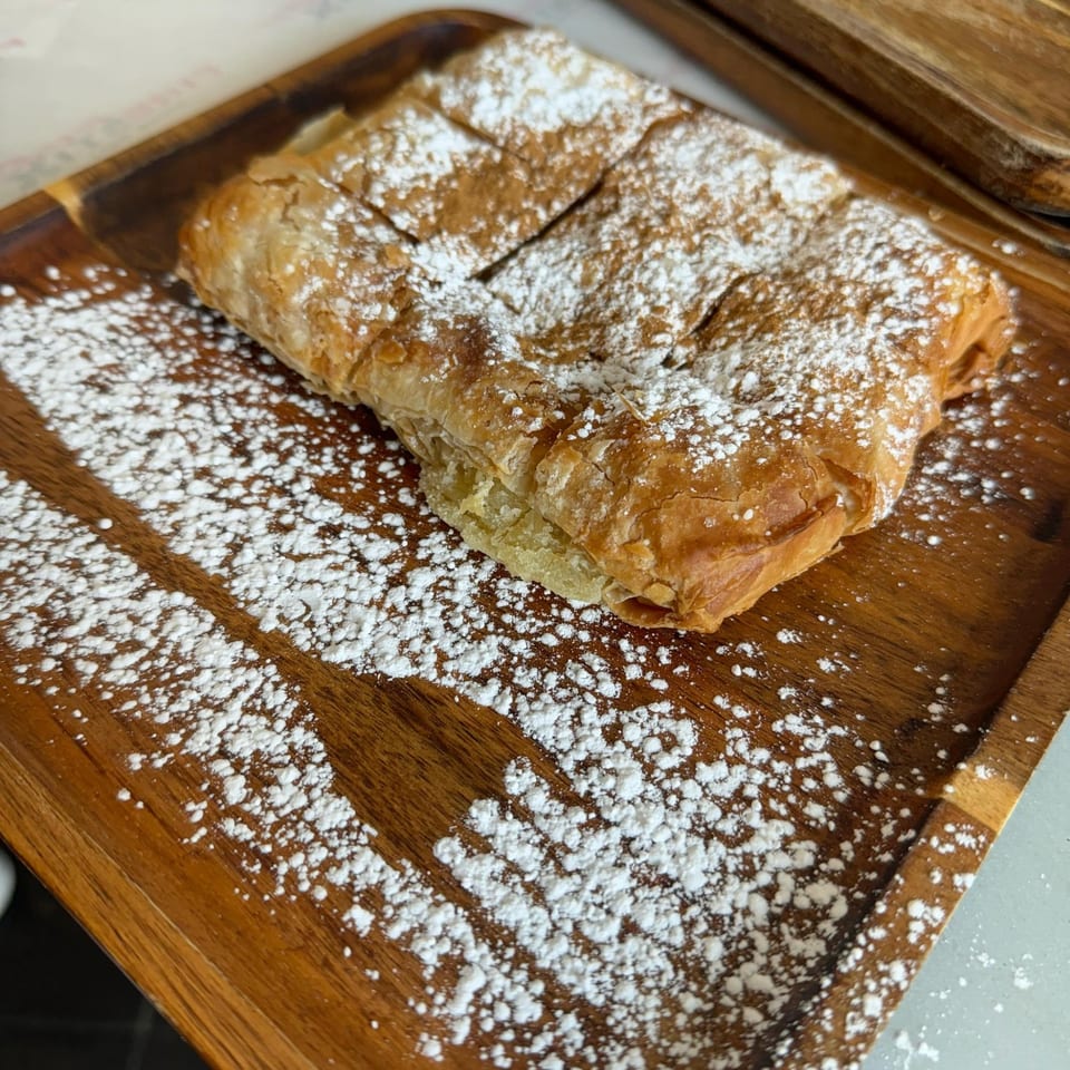 A golden-brown Greek pastry, likely bougatsa, dusted with powdered sugar on a wooden tray.