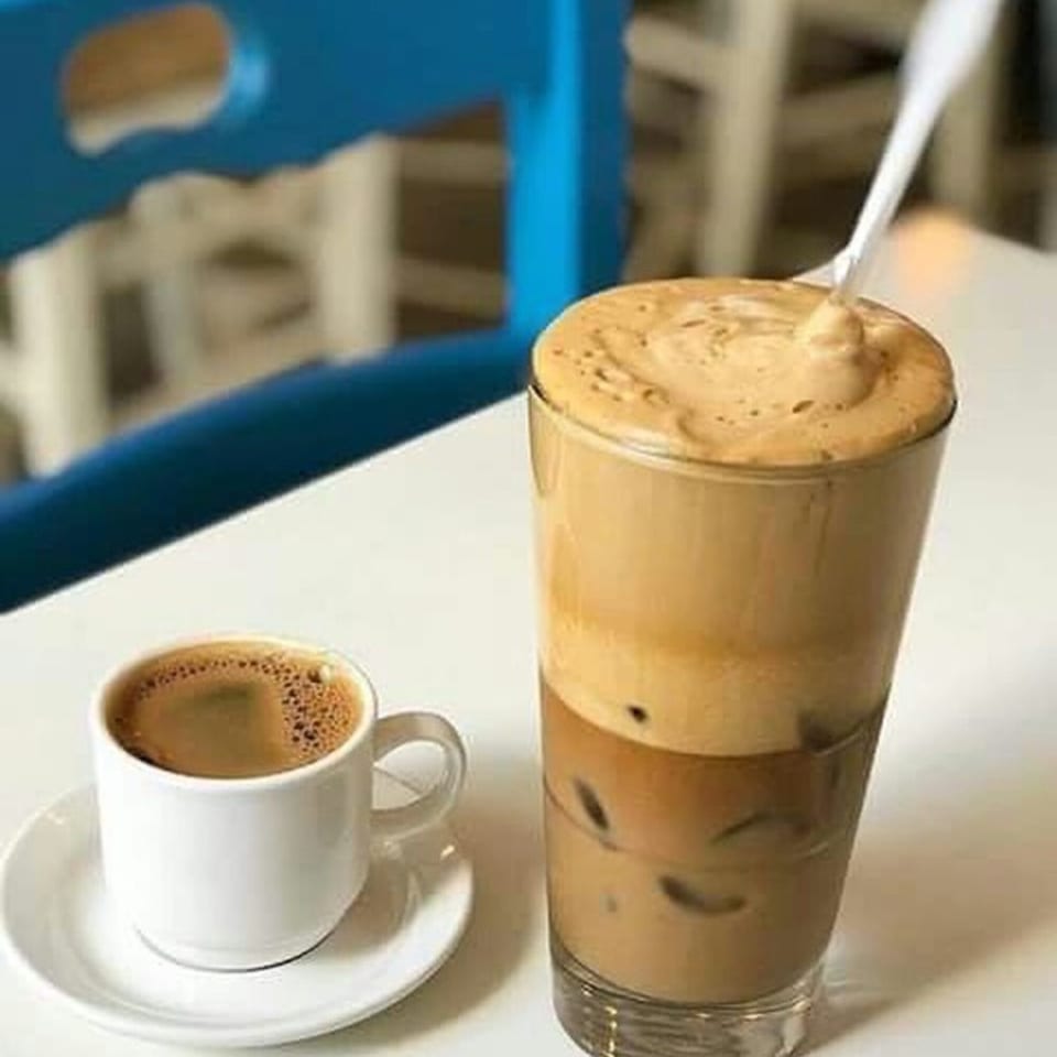 A small white cup of Greek coffee next to a tall glass of iced frappe with a spoon.