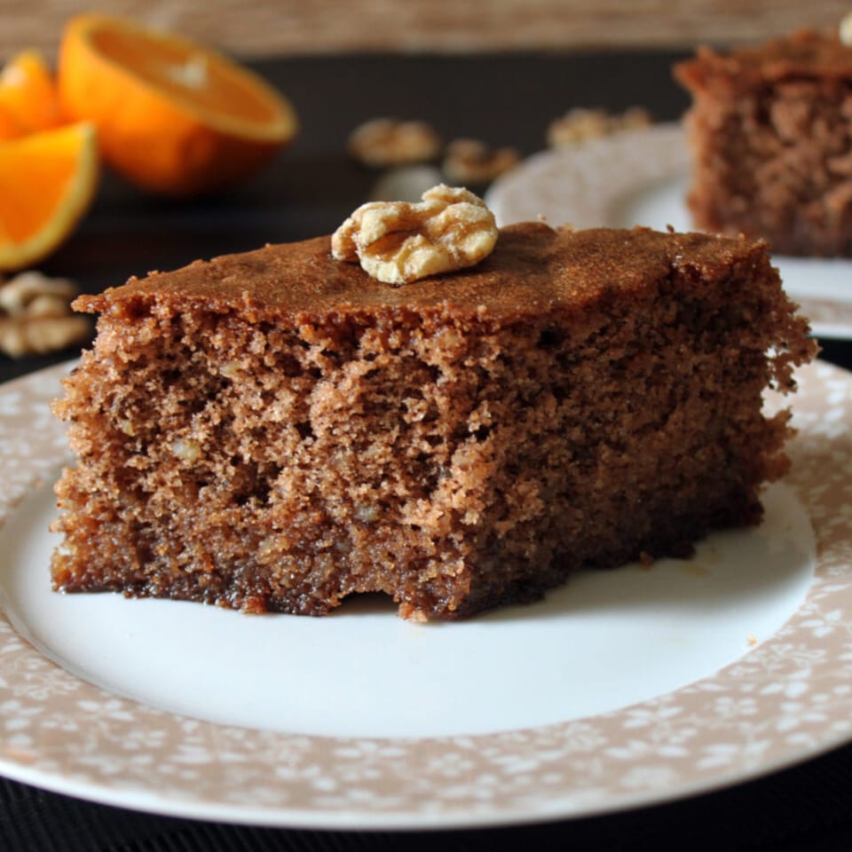 A slice of Greek walnut cake, karidopita, topped with a walnut, on a white plate with oranges.
