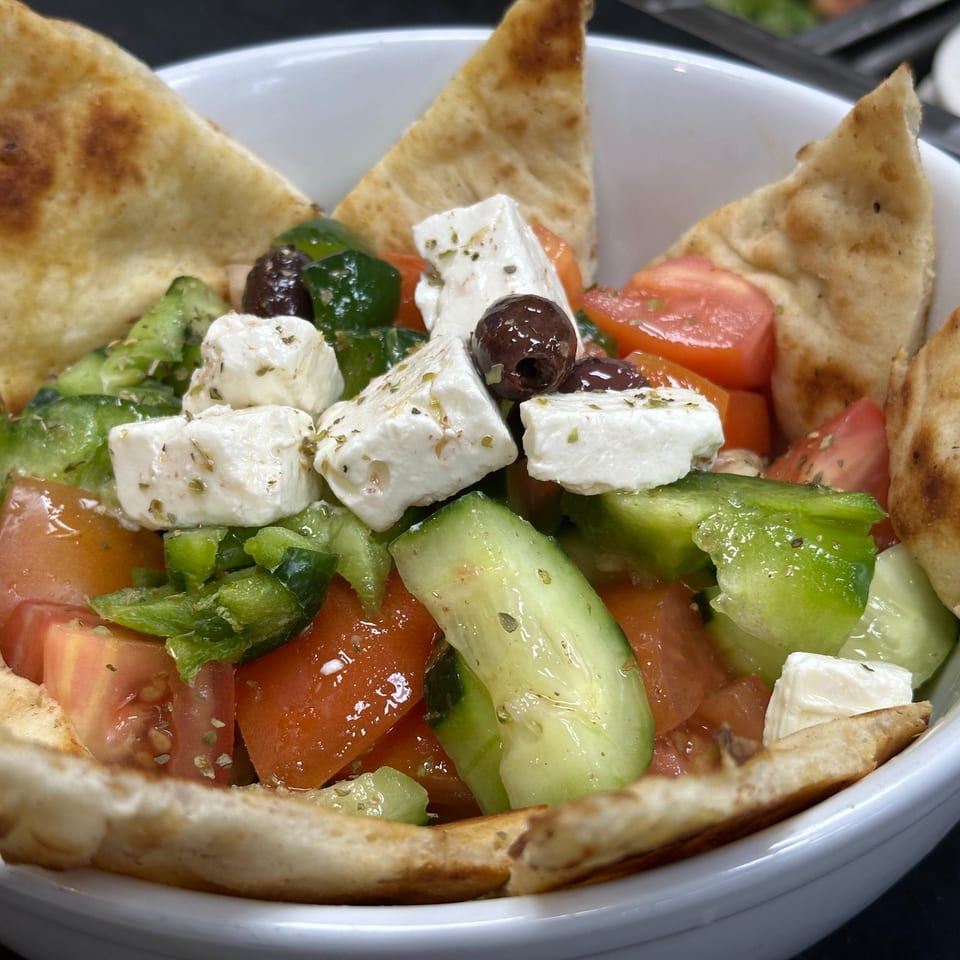 Close-up of a Greek salad with feta, olives, tomatoes, cucumbers, and pita bread in a white bowl.