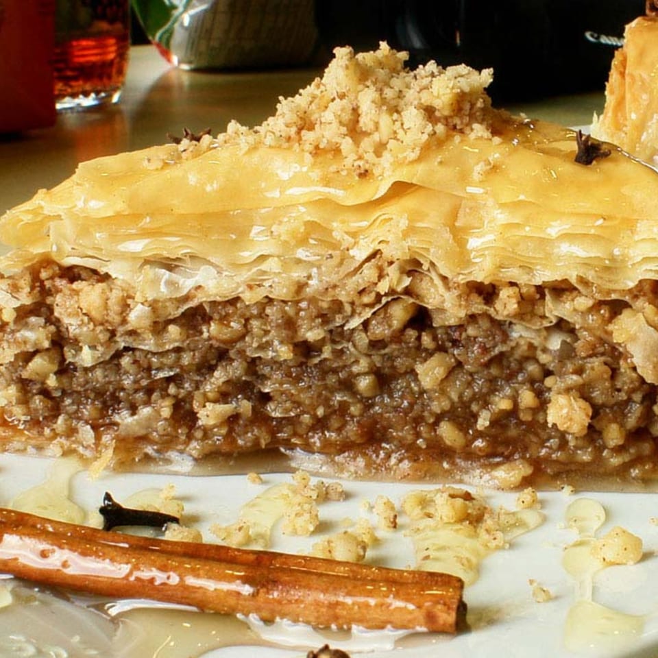 Close-up of a slice of baklava with honey, nuts, and a cinnamon stick on a white plate.