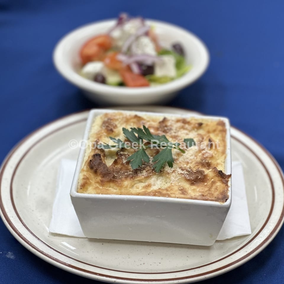 A square dish of golden-brown pastitsio with parsley, and a blurred Greek salad in the background.